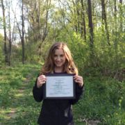 Nicole Thompson standing in a wooded area holding her award certificate